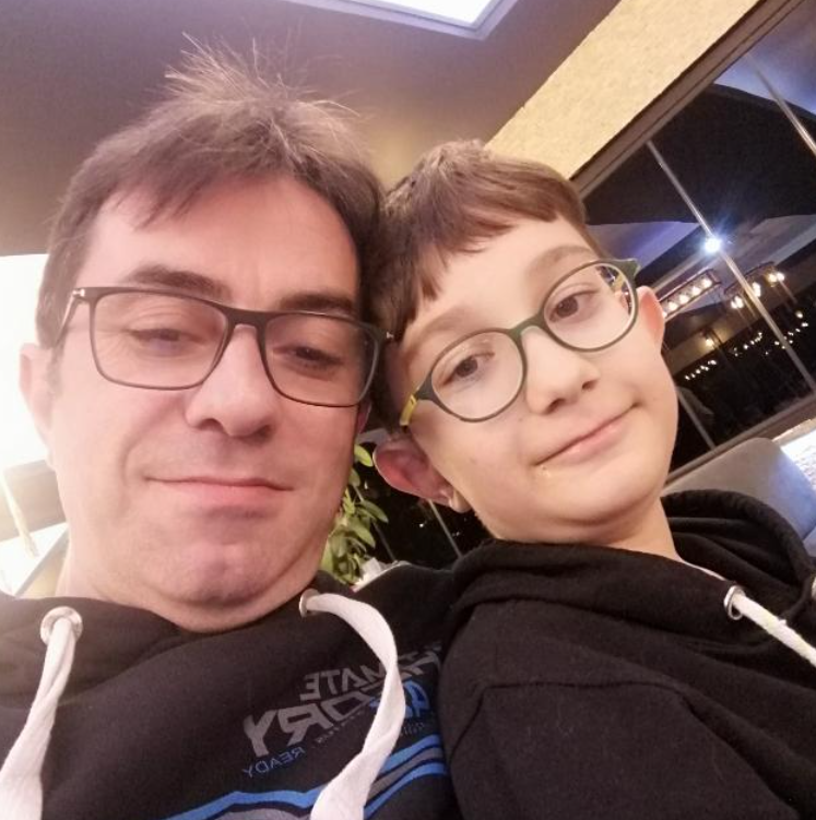 Close-up selfie of a man and a boy wearing glasses, smiling at the camera while leaning their heads together indoors; both in black hoodies.