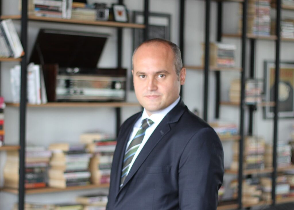 IMG-20160829-WA0005 1 - Rising Stars Spor Aş. Professional man in a dark suit and striped tie standing in a book-filled room, posing for a portrait in an office/library setting.