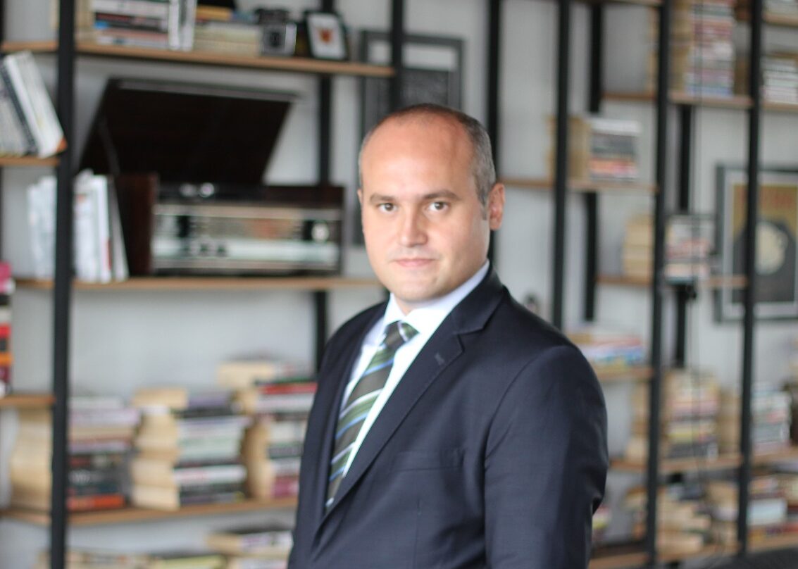 IMG-20160829-WA0005 1 - Rising Stars Spor Aş. Professional man in a dark suit and striped tie standing in a book-filled room, posing for a portrait in an office/library setting.