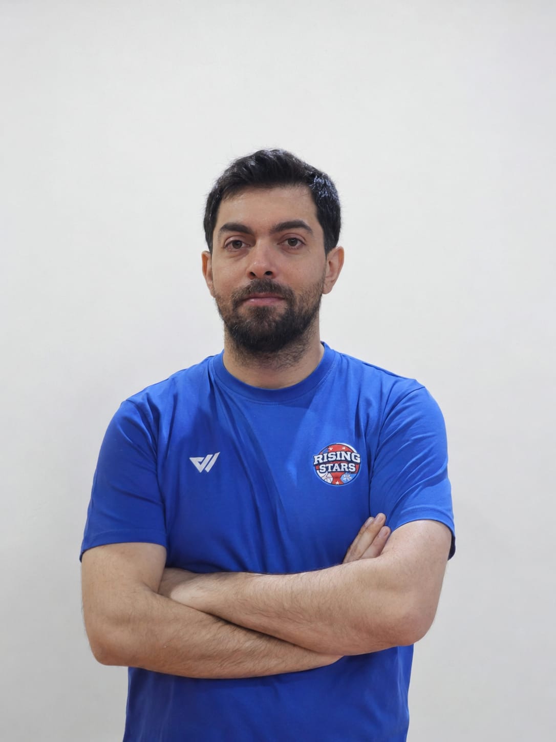 WhatsApp Image 2026-05-17 at 194004 - Rising Stars Spor Aş. Man with dark hair and beard wearing a blue Rising Stars t-shirt, arms crossed, standing against a plain light wall.