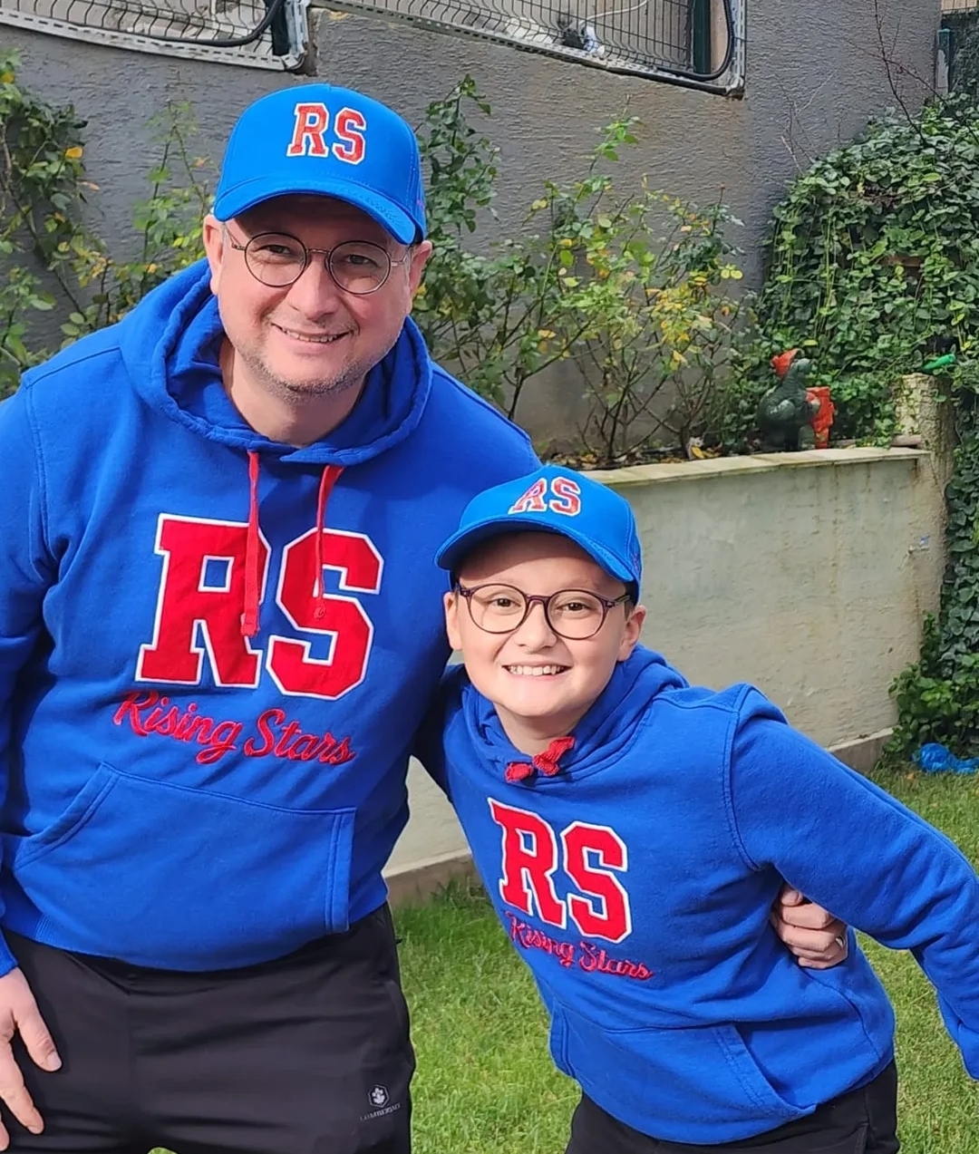 Adult and child in matching blue RS Rising Stars hoodies and caps, smiling together in a backyard garden.