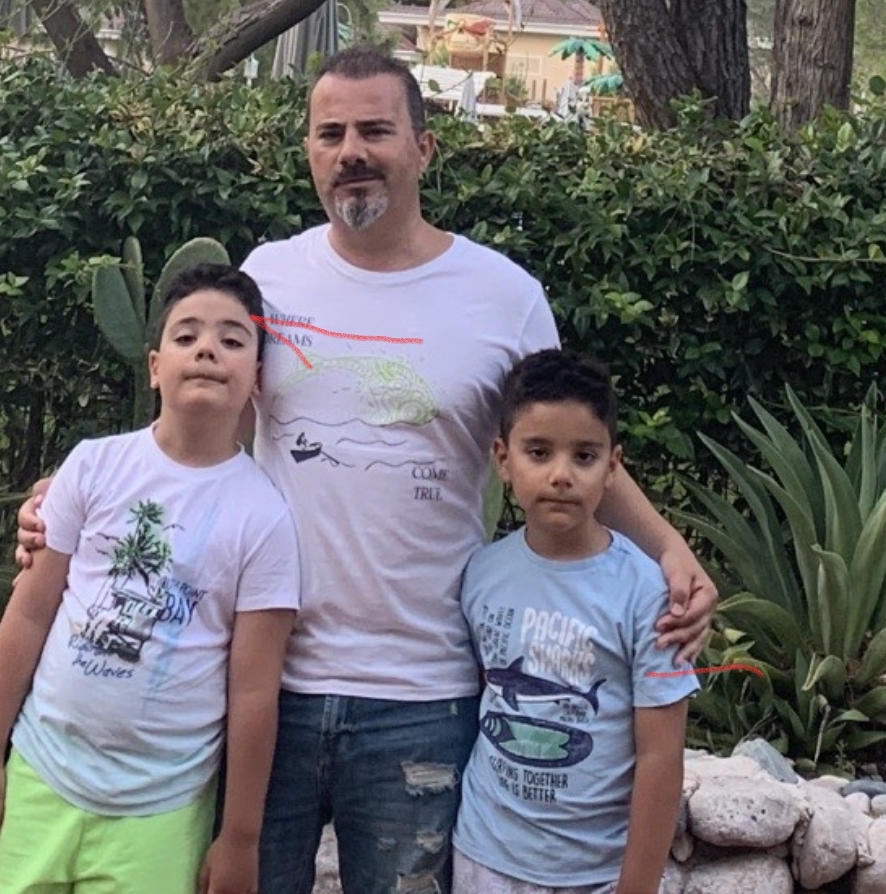 Father and two sons stand together outdoors in a garden, wearing light T-shirts, with greenery and plants in the background.