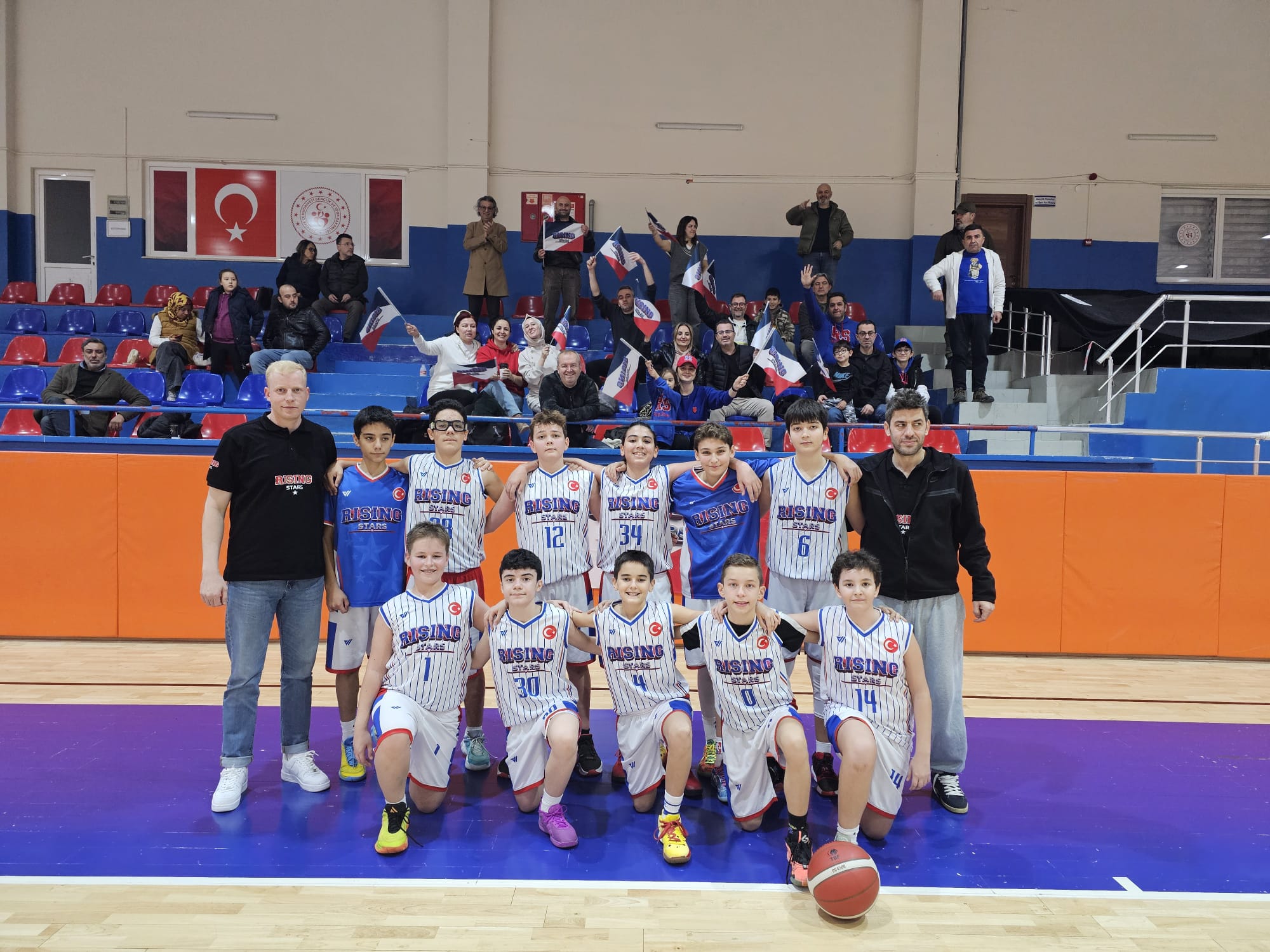 Youth basketball team in white-blue striped jerseys posing for a group photo in a gym, coaches and supporters in the background.