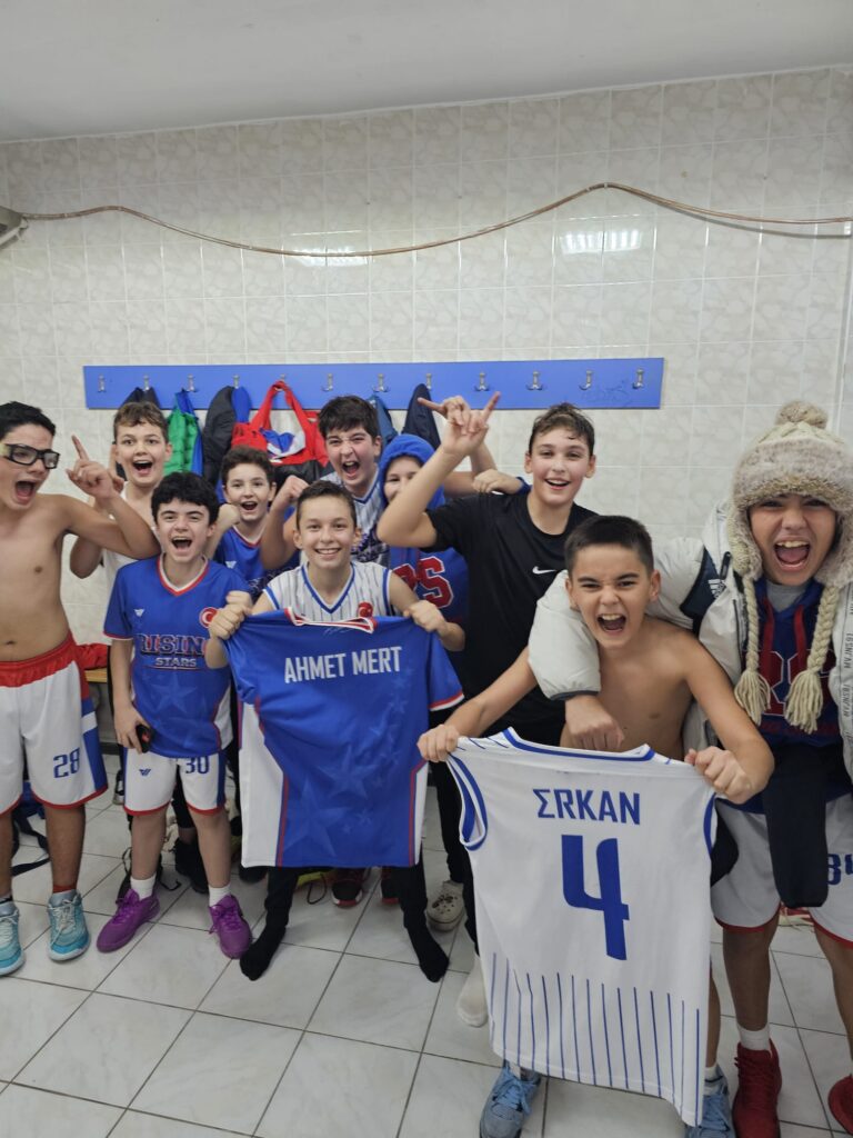 Group of young basketball players in a locker room celebrating, some in blue/white uniforms, holding jerseys and a banner.